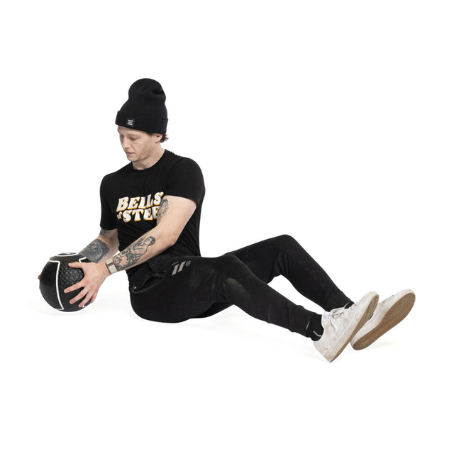 Seated on the floor in black attire and a beanie, this individual holds a Bells of Steel Mighty Grip Medicine Ball with focused intent. Against a plain white backdrop, their white sneakers contrast the intricate tattoos on their arms, embodying fitness dedication with every superior grip.