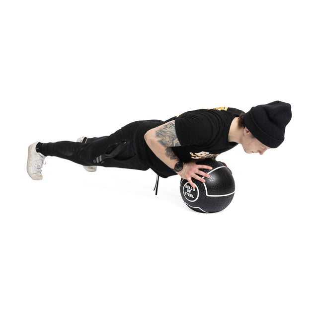 Dressed in black with a beanie, an individual balances in a push-up position on a Bells of Steel Mighty Grip Medicine Ball against a plain white background, highlighting the product's superior grip and their impressive fitness.