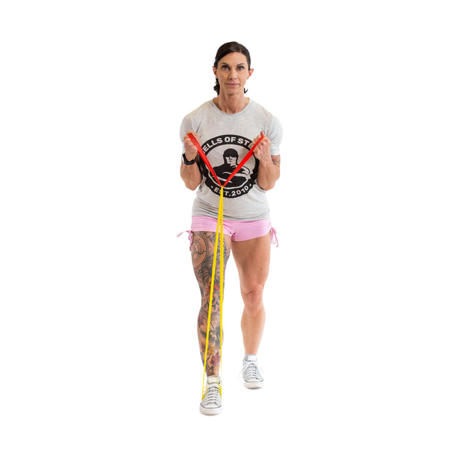 A person in a white T-shirt and pink shorts is working out with the yellow Bells of Steel Mini Band, crafted from high-grade latex. They are standing on it with one foot while pulling it with both hands. The individual, wearing white sneakers, has tattoos on one leg.