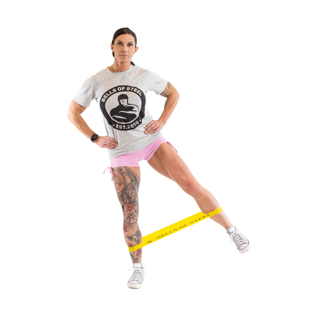 A person works out with a yellow Mini Band from Bells of Steel, crafted from high-grade latex, wrapped around their legs. They're wearing a gray "Bells of Steel" t-shirt, paired with pink shorts and sneakers. As they confidently lift one leg to the side, their hands rest on their hips.