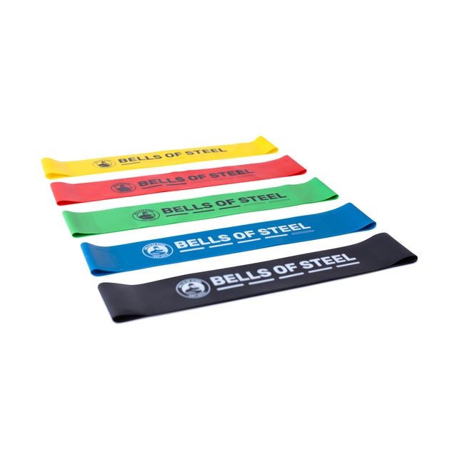 Five vibrant Mini Bands from Bells of Steel, crafted from high-grade latex in yellow, red, green, blue, and black, are arranged in an upward diagonal on a plain white background.