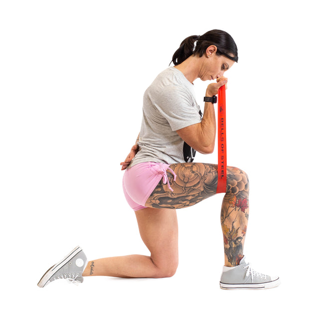 A person kneels on one knee, gripping a Mini Band from Bells of Steel, crafted from high-grade latex, over their shoulder. They wear a gray shirt, pink shorts, and gray sneakers. Their tattooed leg adds a personal touch against the plain white background.