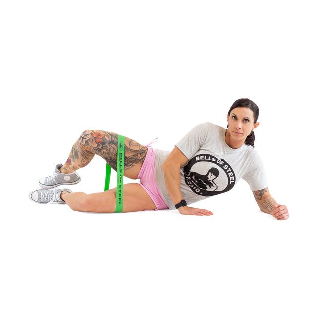 A tattooed individual is lying on their side, performing a leg exercise with a Bells of Steel Mini Band made from high-grade latex. They are wearing a grey T-shirt, pink shorts, and grey sneakers against a clean white backdrop.