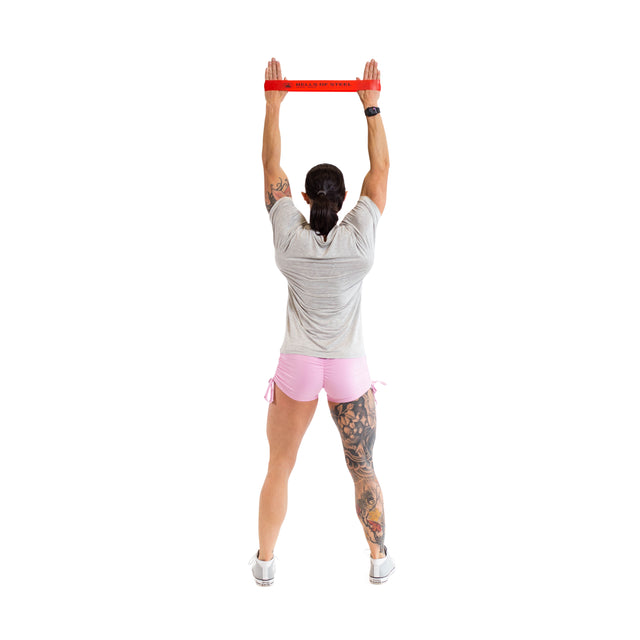 An individual, dressed in a gray shirt, pink shorts, and white sneakers, stands with their back to the viewer while stretching a high-grade red resistance band overhead. They have a tattoo on one leg. The white background highlights the adaptability of Bells of Steel's Mini Bands in any setting.