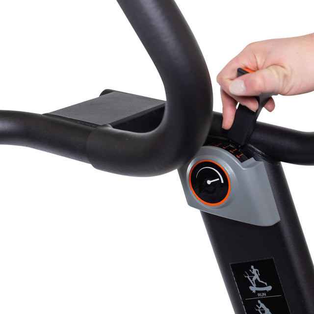 A close-up shows a hand adjusting the magnetic resistance on the Blitz Manual Treadmill by Bells of Steel. The hand turns a knob near the curved black handlebars, ideal for cardio workouts, while the control panel features a running stick figure icon.