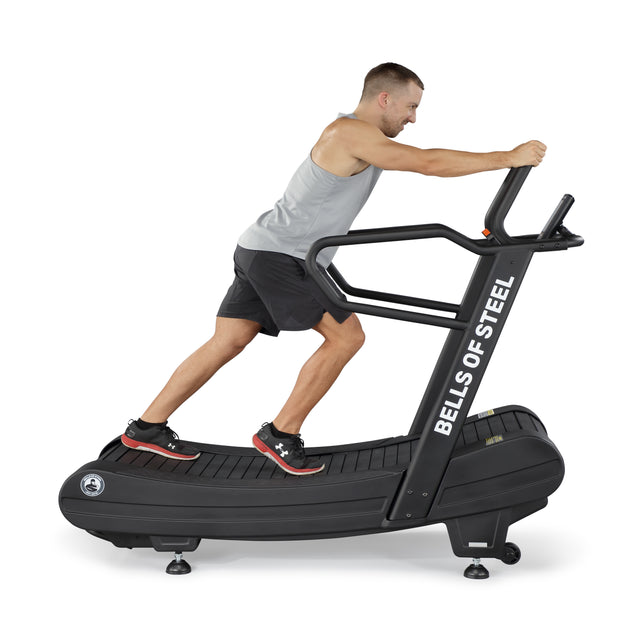 A person is running on the Blitz Magnetic Resistance Manual Treadmill by Bells of Steel, perfect for cardio. They wear a gray sleeveless shirt, black shorts, and sneakers. The treadmill is black and set against a white background.