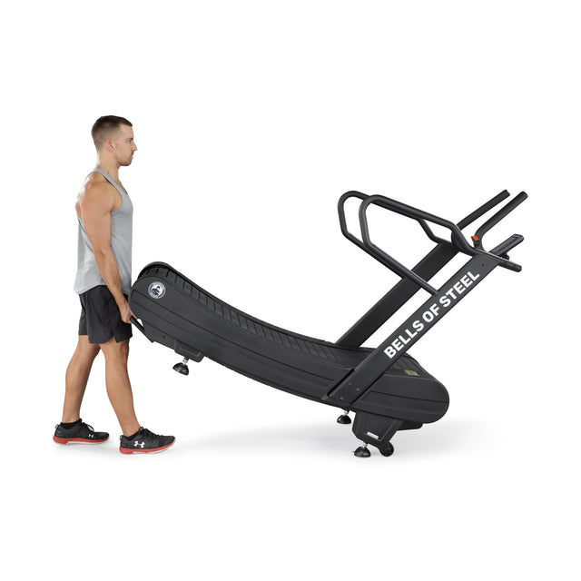 A man in athletic wear tilts a "Bells of Steel" Blitz Magnetic Resistance Manual Treadmill onto its wheels, showing its portability for cardio workouts. The compact black treadmill with curved handrails offers magnetic resistance for an enhanced exercise experience.