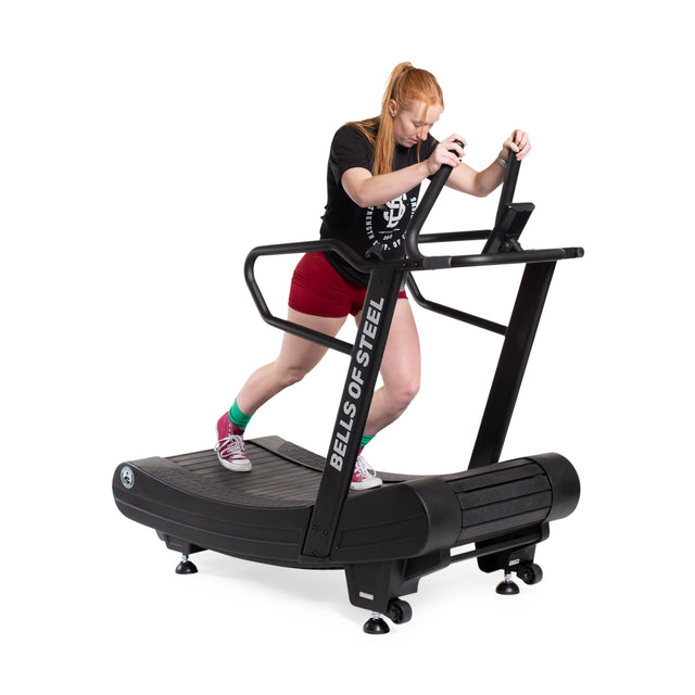 Wearing a black T-shirt, red shorts, green socks, and red sneakers, a person with long hair does cardio on the Blitz Magnetic Resistance Manual Treadmill by Bells of Steel. They grip the handles firmly while concentrating on the display.