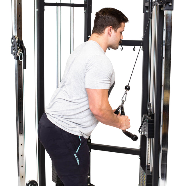 A man wearing a gray t-shirt and black shorts is exercising on a cable machine, performing tricep pushdowns using the Bells of Steel MULTI-PURPOSE V STYLE BAR. The gym environment features metal bars and pulleys, complemented by rubberized grips for safe cable training.