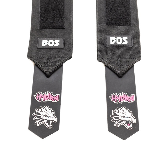 The Mighty Wrist Wraps from Bells of Steel are competition-style wrist wraps featuring two black straps, each displaying the "BOS" logo near the top and a stylized dragon skull with "HYDRA" in pink text underneath. Designed as powerlifting essentials, these wraps come equipped with Velcro closures and highlight a sleek design.