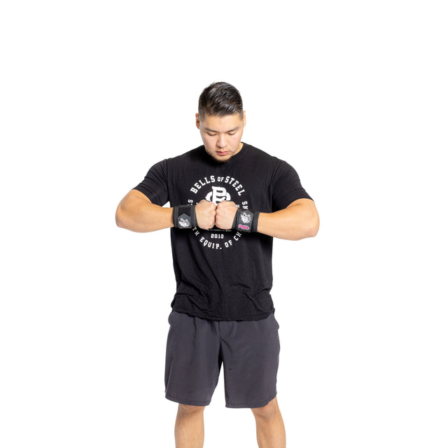 A person wearing a black "Bells of Steel" shirt and gray shorts stands against a white background, looking down with hands on wrists. The vibrant "Mighty Wrist Wraps," competition-style wraps from Bells of Steel and considered powerlifting essentials, highlight their fitness gear.