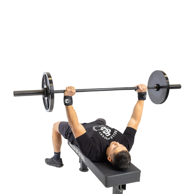 A person performs a bench press on a flat bench, lifting a barbell with weights. Dressed in powerlifting essentials, they sport a black shirt, shorts, and sturdy Mighty Wrist Wraps by Bells of Steel. The white background emphasizes the exercise.