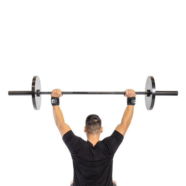 A person in a black shirt lifts a barbell overhead while standing against a plain white background, showcasing a well-defined physique. They wear Bells of Steel's Mighty Wrist Wraps for added stability.