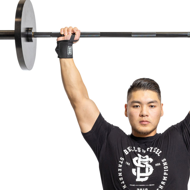 A man wearing a black shirt from "Bells of Steel" is seen holding a barbell overhead with one hand, displaying powerlifting essentials. His other arm, bent at his side, is wrapped in "Mighty Wrist Wraps" designed for competition style. The background is plain white.