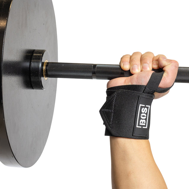 A person lifts a barbell with large black weight plates, using Bells of Steel's Mighty Wrist Wraps on their right wrist. The strap sports a white "BOS" logo against its black fabric. The background is pristine white, emphasizing the intensity of the lift.