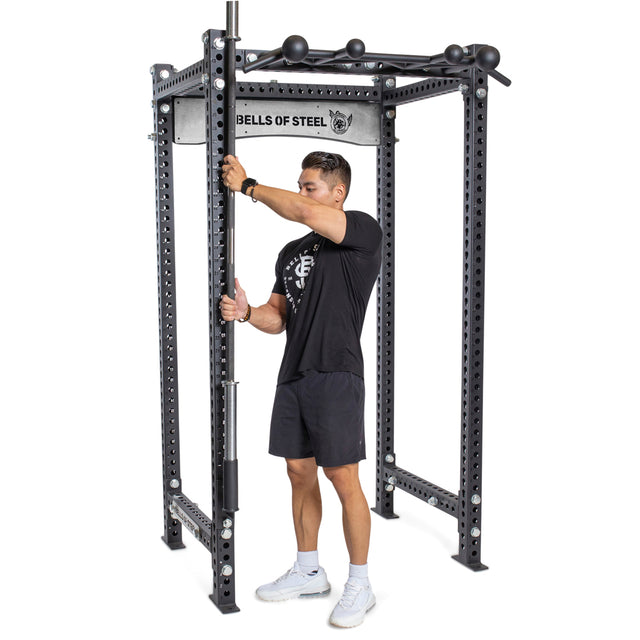 A person dressed in a black workout outfit is adjusting a barbell on a Bells of Steel power rack, which is equipped with the Vertical Mount Barbell Holder Rack Attachment for easy home gym organization. The metal rack includes multiple holes for adjustments, and the floor beneath it is white, complementing the person's matching white shoes.