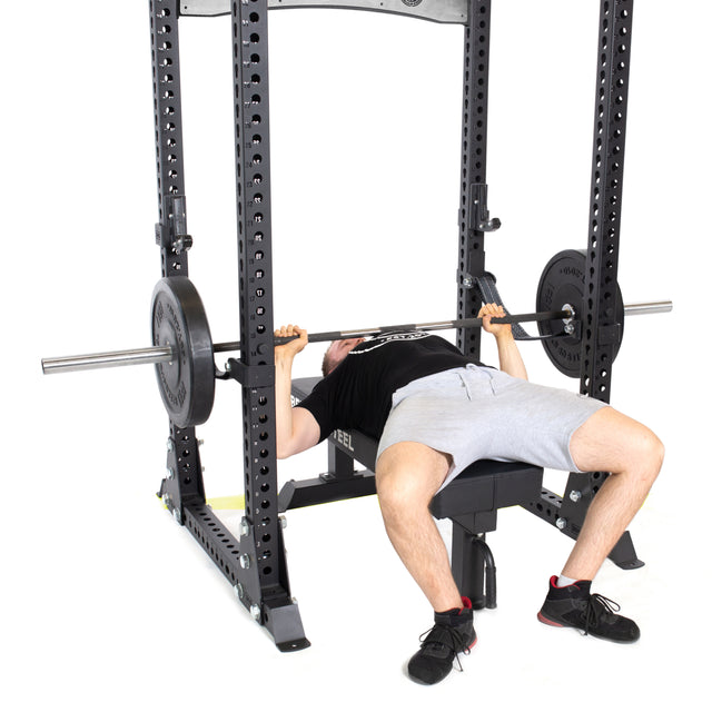 Using the Bells of Steel Manticore Flat Foot Power Rack Builder (3" x 3", 1" Holes), a person is bench pressing with a barbell that has a 1,000-pound capacity. They are lying on a bench, lifting the weighted barbell above their chest while wearing a black shirt and gray shorts.