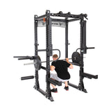 A person is squatting inside the Bells of Steel Manticore Flat Foot Power Rack Builder (3" x 3", 1" Holes) with a barbell loaded with weights. The open ecosystem of the rack includes various attachments, and the individual is dressed in a black shirt, white shorts, and sneakers.