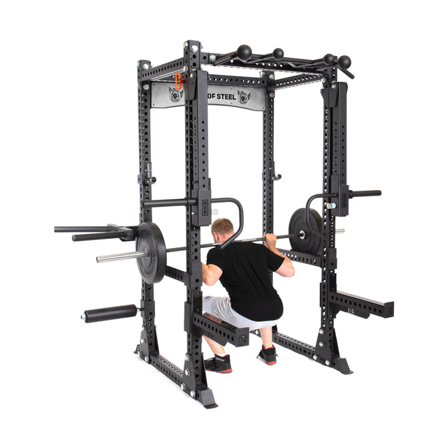 A person is squatting inside the Bells of Steel Manticore Flat Foot Power Rack Builder (3" x 3", 1" Holes) with a barbell loaded with weights. The open ecosystem of the rack includes various attachments, and the individual is dressed in a black shirt, white shorts, and sneakers.