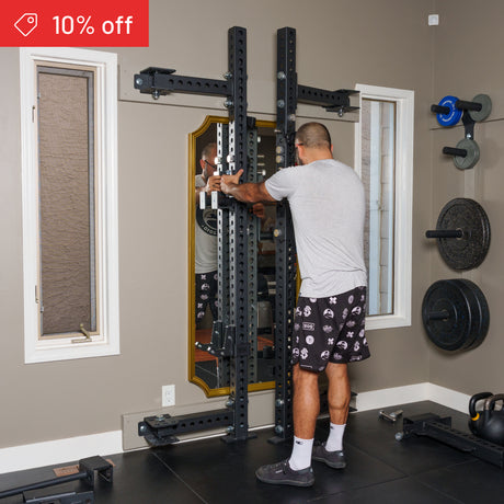A man in a home gym adjusts the Bells of Steel Manticore Folding Half Rack Builder (3" x 3", 1" holes) mounted to the wall. He wears a gray tee, patterned shorts, and sneakers. Weight plates are nearby. A "10% off" sale tag is shown.