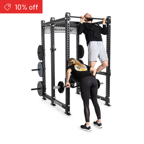 A man does a pull-up while a woman lifts a barbell on the Bells of Steel Manticore Six Post Power Rack Builder (3" x 3", 1" Holes), with weight plates stored on the side. A red "10% off" banner is in the corner.
