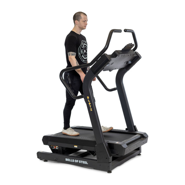 A person stands on a stationary Blitz Mountain Climber Treadmill, featuring FTMS Bluetooth connectivity and displaying "Bells of Steel." Wearing a black T-shirt and dark pants, they are ready to tackle workouts using the treadmill’s advanced screen and handlebars.