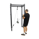 A person in a black shirt and shorts uses the Bells of Steel Tricep Rope Extension at the gym, with a metal frame and weights on pulleys, standing on a platform for focused tricep targeting.