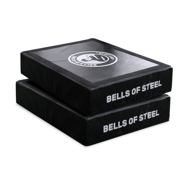 Two stacked, rectangular, black crash pads called the Hydra Nordic Curl Package by Bells of Steel feature "BELLS OF STEEL" in white text. The top pad includes a circular logo. These pads are perfect for minimizing noise and impact during weightlifting or Nordic hamstring curls, providing stability and comfort in your workout routine.