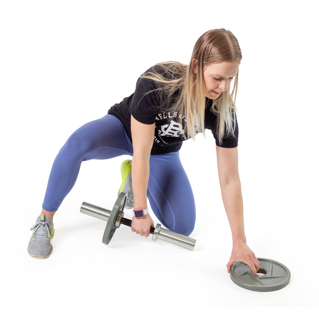 A person kneels on one knee, gripping a Bells of Steel Loadable Dumbbell Handle with a weight plate on one end. Reaching for another plate on the floor, they're dressed in a black shirt, blue leggings, and gray sneakers. The scene is set against a white background showcasing gym equipment essentials.