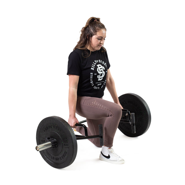 A woman dressed in a black T-shirt and brown leggings expertly performs a half-kneeling deadlift with an Open Trap Bar / Hex Bar from Bells of Steel on a white background. Wearing white sneakers, she gracefully showcases her technique, emphasizing the barbell's rotating sleeves for seamless motion.