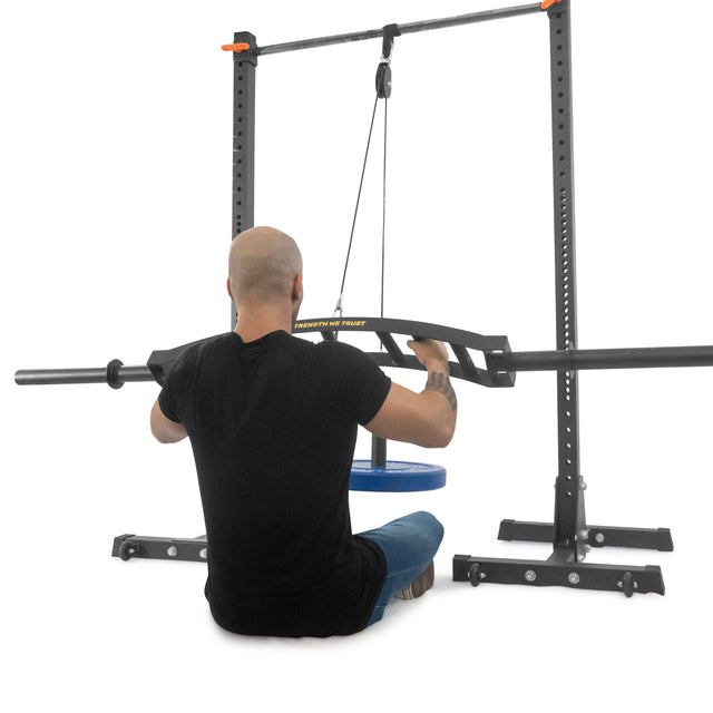 A bald man in a black T-shirt and jeans uses an Omni Bar from Bells of Steel, attached to weighted plates, in a minimalist gym. Power racks and meticulously knurled equipment emphasize his strength-focused workout routine.