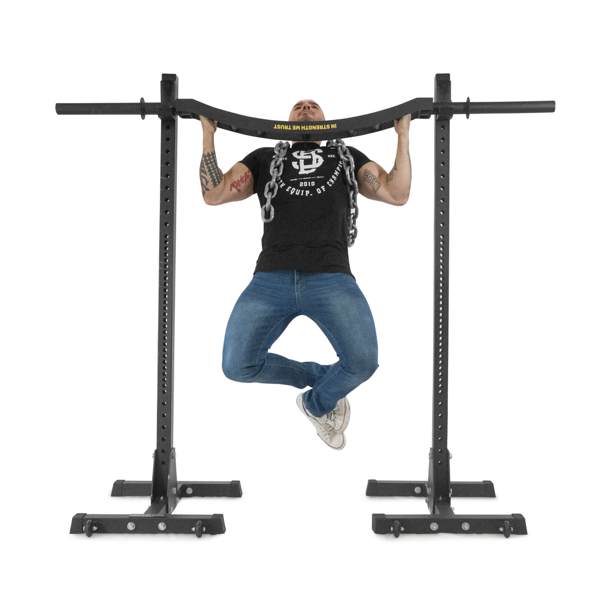 A person in a black t-shirt and blue jeans is doing a pull-up with a neutral grip on a sleek power rack. Their arms have tattoos, and they wear white sneakers. The well-lit exercise space features the distinct knurling of the Bells of Steel Omni Bar.