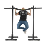 A person in a black t-shirt and blue jeans is doing a pull-up with a neutral grip on a sleek power rack. Their arms have tattoos, and they wear white sneakers. The well-lit exercise space features the distinct knurling of the Bells of Steel Omni Bar.