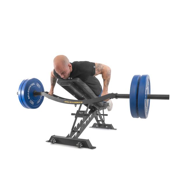 In a studio setup, someone with a shaved head and arm tattoos performs back extensions on a Roman chair using blue Omni Bar weights by Bells of Steel. They're wearing a black shirt, surrounded by power racks against a white background.