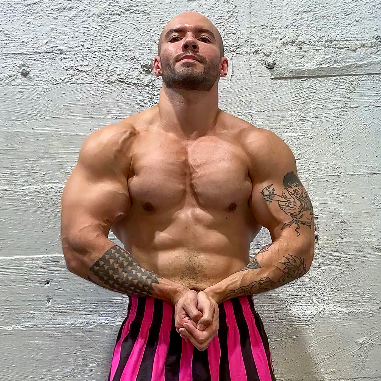 A muscular man poses against a textured wall, flaunting tattoos on his arms and exuding the strength of Bells of Steel power racks. He confidently sports pink and black striped pants, echoing the gritty elegance reminiscent of an Omni Bar's knurling.