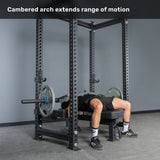 Lying on a bench with an arched back, a person grips the Bells of Steel Arch Nemesis Swiss Bar in a power rack. Text above reads: "Cambered arch extends range of motion." This specialty bar boosts pressing performance.