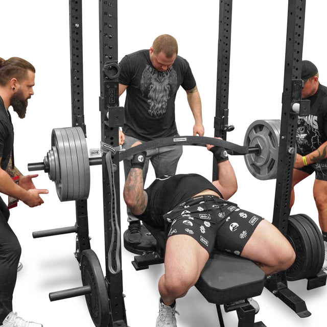 Using the Bells of Steel Arch Nemesis Swiss Bar, a person executes a heavy bench press in a gym with three spotters, their focus sharp against the white background and contrasting black specialty barbells.