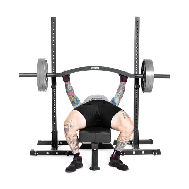 A tattooed individual bench presses using a Bells of Steel Arch Nemesis Swiss Bar on a weight bench, lying back and holding the loaded specialty barbell in a gym setting.