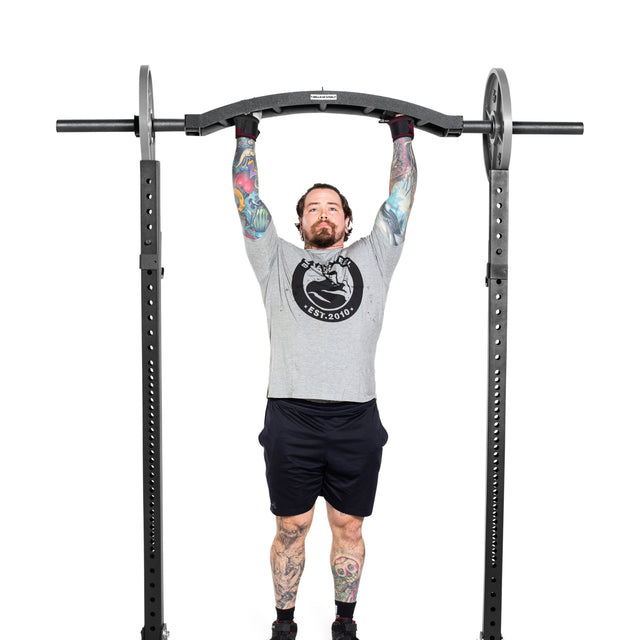 In a gym, a tattooed person in a gray shirt and black shorts performs a pull-up on a squat rack equipped with the Bells of Steel Arch Nemesis Swiss Bar.