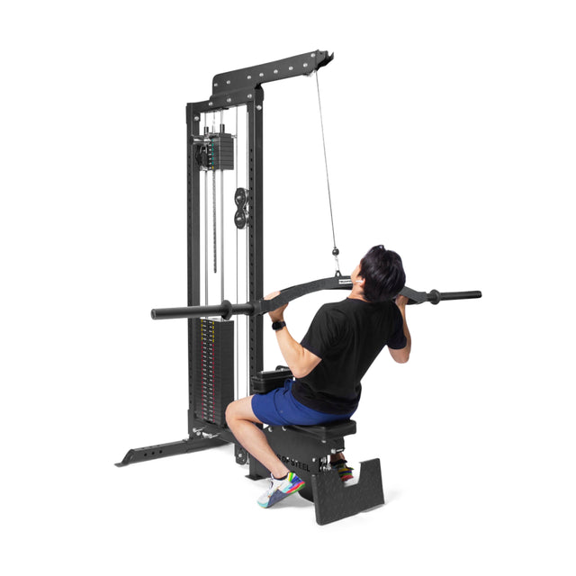 A person in a black shirt and blue shorts performs a lat pulldown on the cable machine. Gripping the Arch Nemesis Swiss Bar by Bells of Steel with both hands, they focus on maximizing muscle engagement while a stack of weights awaits their next set.