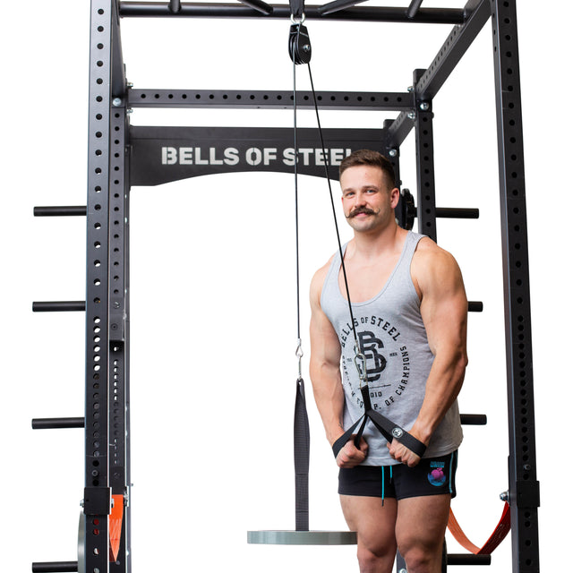 A man wearing a tank top and shorts is using the Bells of Steel Cable Pulley to perform triceps pushdowns in the gym. Positioned against a white background, he sports a mustache and concentrates on his workout, gripping the cable attachment with both hands.