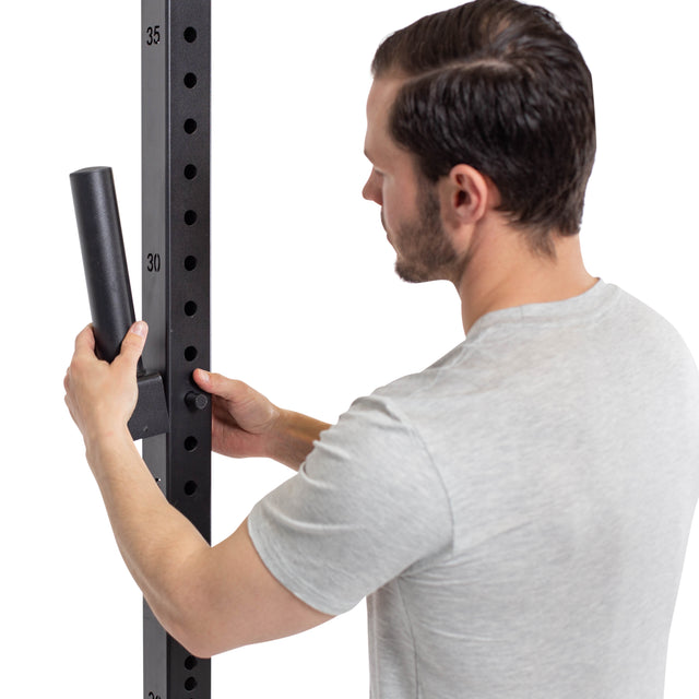 A man in a gray t-shirt adjusts the Bells of Steel Pin Plate Pegs on a tall, perforated metal structure, possibly a power rack, with plate pegs and weight plates indicating a fitness or exercise context.