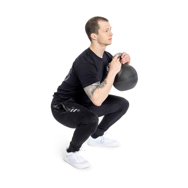 Clad in black, a person expertly squats with the Bells of Steel adjustable kettlebell featuring a plastic bottom. The kettlebell's flexible weight increments are managed close to their chest, while they stand on a white background wearing light-colored sneakers.