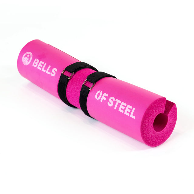 A vivid pink foam "Bells of Steel" barbell pad with black straps, ergonomically crafted for comfort, is rolled up to display its color and branding on a white background.