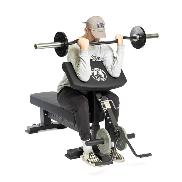 A person wearing a gray cap and sweatshirt is stylishly performing a bicep curl on a seated preacher curl machine, equipped with Bells of Steel Bench Attachments. In their checkered shoes, they are lifting a barbell with weights in a gym setting.