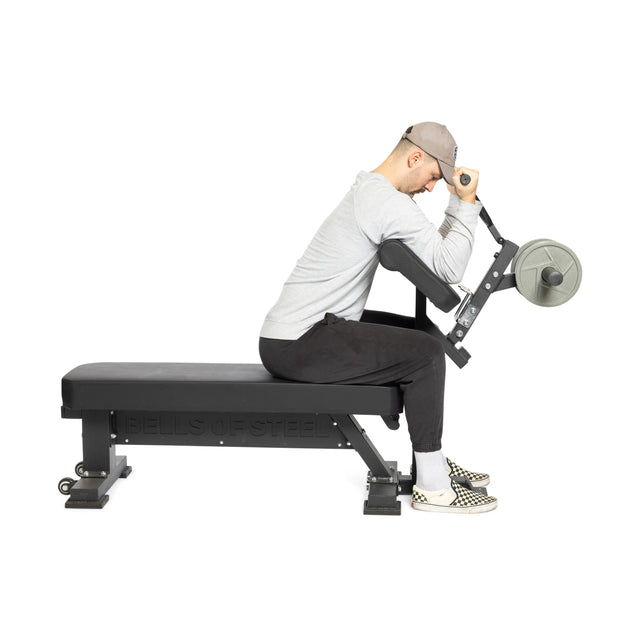 A person dressed in athletic wear is seated on a Bench Attachment from Bells of Steel, performing a preacher curl with a weight-loaded barbell. The individual appears focused, against a neutral background that highlights the exercise.