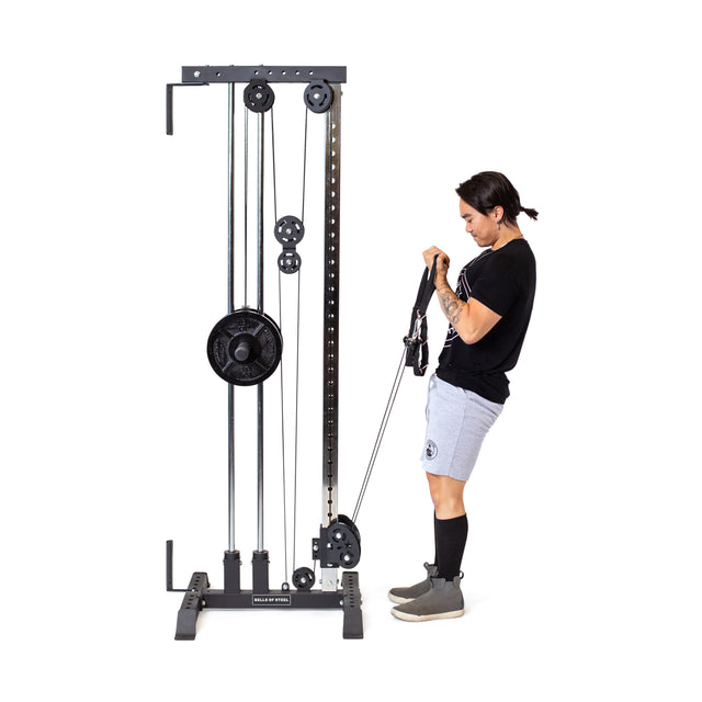 In their home gym, a person uses the Bells of Steel Cable Tower for bicep curls. They sport a black shirt, gray shorts and sneakers, and black socks. The Cable Tower offers versatile exercises with multiple pulleys and weight plates.