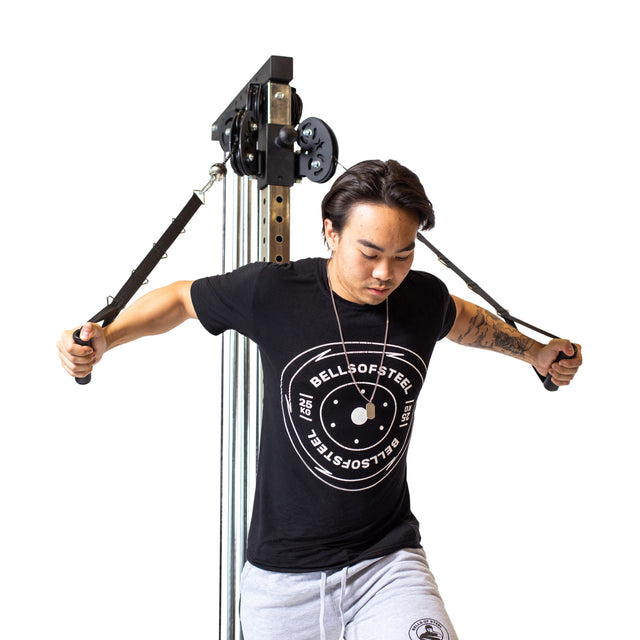 Someone in a black T-shirt and grey sweatpants uses the Bells of Steel Cable Tower for chest exercises, expertly pulling the cables outward. The workout takes place in a sleek home gym with a clean, white background.