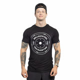 Wearing a "Bamboo T-Shirt" from Bells of Steel, featuring a circular design similar to a weight plate, an individual stands confidently. Tattoos adorn both arms and they sport a backwards black cap, effortlessly exuding coolness.