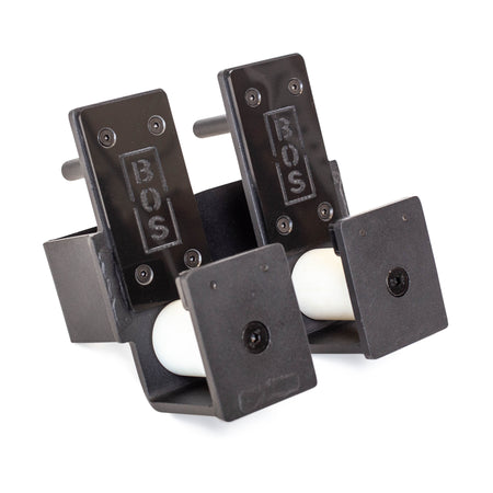 The Roller J-Cups from Bells of Steel are a pair of black foot pedals featuring sturdy and durable nylon rollers with a white spherical design underneath. Embossed with "BOS," their simple, industrial aesthetic complements any setting. Displayed against a plain white background, they stand out as essential accessories.